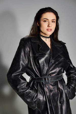 A stunning plus size woman exudes confidence in a black leather trench coat on a neutral grey backdrop.