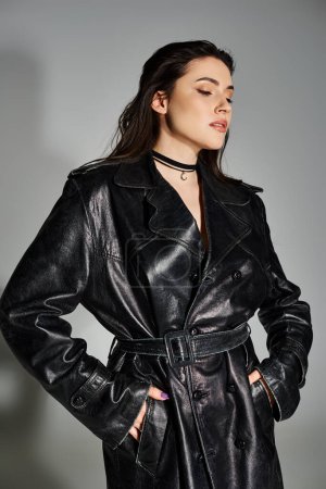 A stunning plus size woman striking a pose in a black leather trench coat against a gray backdrop.