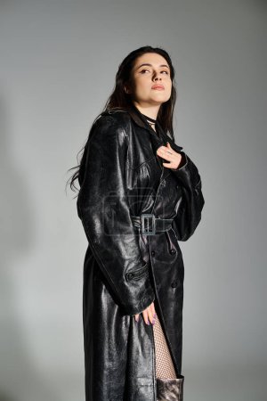 Eine schöne Plus-Size-Frau posiert selbstbewusst in schwarzem Mantel und Netzstrümpfen vor einem glatten grauen Hintergrund.