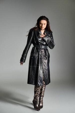 Mujer de talla grande en abrigo de cuero negro de pie sobre un fondo gris, enfocado en su teléfono celular.