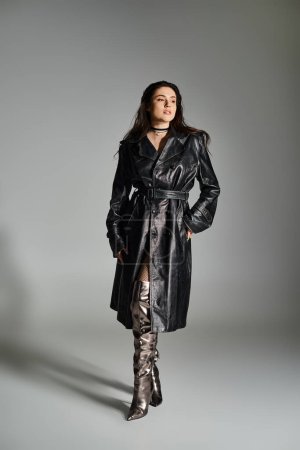 Eine atemberaubende Plus-Size-Frau posiert in stylischem schwarzen Mantel und Stiefeln vor grauem Hintergrund.