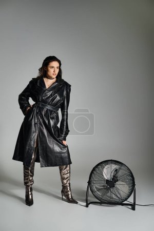 Photo for Stunning plus size lady in chic black coat and boots standing gracefully next to a retro fan on gray background. - Royalty Free Image