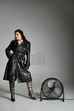 Foto de Una impresionante mujer de talla grande con un elegante abrigo negro y botas de pie con gracia junto a un ventilador vintage en un fondo gris. - Imagen libre de derechos