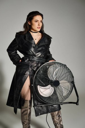 A beautiful, plus size woman strikes a pose in a stylish trench coat, holding a fan against a gray backdrop.