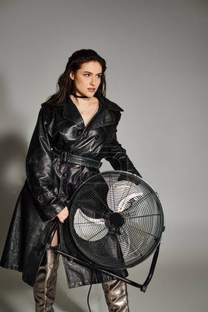 A beautiful plus size woman posing in a fashionable leather coat, holding a fan, exuding confidence and style on a gray backdrop.