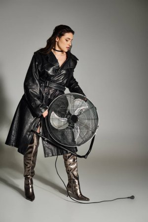 Une belle femme de taille plus dans un manteau noir pose gracieusement, tenant un ventilateur délicat sur un fond gris.