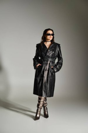 A beautiful plus size woman exudes confidence in a black trench coat and boots against a gray backdrop.