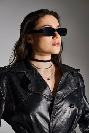 Eine schöne Plus-Size-Frau posiert selbstbewusst in schwarzer Lederjacke und stylischer Sonnenbrille vor grauem Hintergrund.