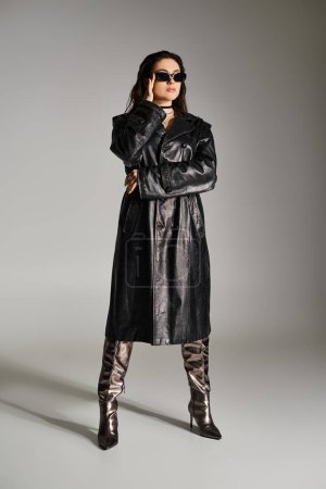Eine Plus-Size-Frau strahlt in stylischem schwarzen Ledermantel und Stiefeln vor grauem Hintergrund Zuversicht aus.