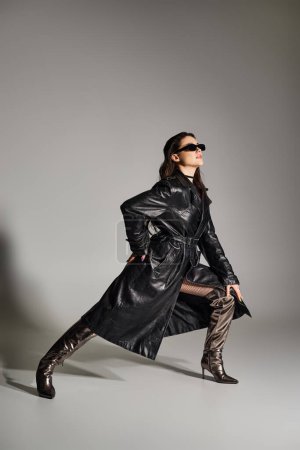 Una hermosa mujer de talla grande posa con confianza en un abrigo negro y botas contra un telón de fondo gris, exudando estilo y gracia.
