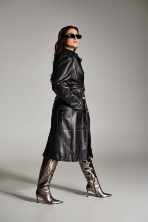 Elegante mujer de talla grande pavoneándose con confianza en un abrigo de cuero negro y botas contra un fondo gris llamativo.