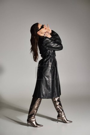 Curvy woman exudes confidence in a black leather coat and boots against a gray backdrop.