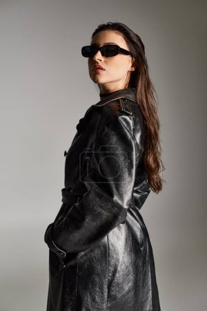 A plus size woman exudes style in a black coat and sunglasses against a gray backdrop, striking a confident pose.