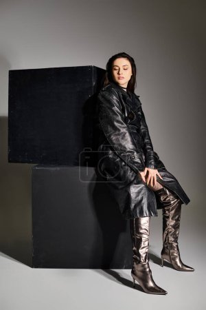 Eine Plus-Size-Frau in stylischer Kleidung sitzt anmutig auf einer schwarzen Schachtel vor grauem Hintergrund.