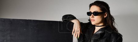 Foto de A stunning plus size woman exudes confidence as she poses in a black leather jacket and stylish sunglasses against a gray backdrop. - Imagen libre de derechos