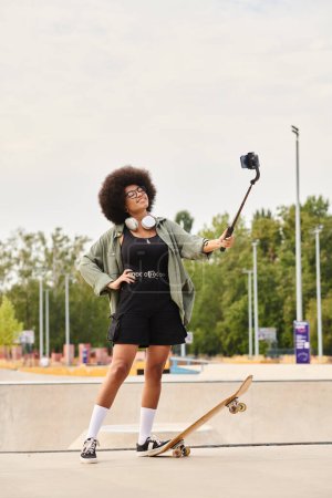 A young African American woman with curly hair stands next to a skateboard, holding a selfie stick in a skate park.