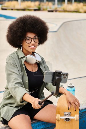 Joven mujer afroamericana con el pelo rizado sentado en una cornisa, sosteniendo un palo selfie en un parque de skate vibrante.