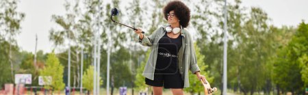 A young African American woman with curly hair confidently holds a selfie stick in an outdoor setting.