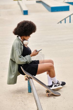 Junge Afroamerikanerin mit lockigem Haar sitzt auf einem Skateboard und benutzt ein Handy in einem belebten Skatepark.