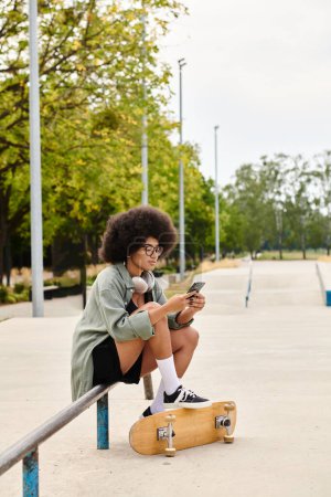 A young African American woman with curly hair sits on a skateboard, engrossed in her cellphone at an outdoor skate park.