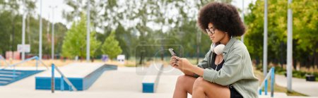A young woman with curly hair sits on a bench, engrossed in her cell phone at a skate park.