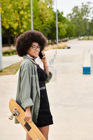 Eine junge Afroamerikanerin mit lockigem Haar multifunktional, indem sie ein Skateboard in der Hand hält und in einem Skatepark mit einem Handy spricht.