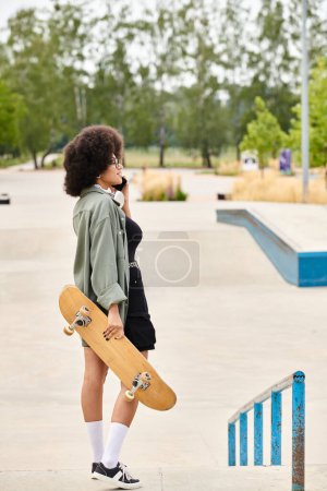 Young African American woman with curly hair, holding a skateboard while chatting on a cell phone in a vibrant outdoor skate park.
