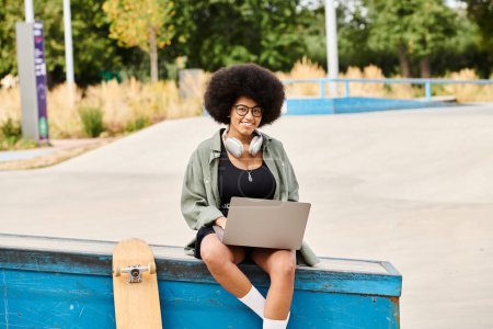 A black woman with curly hair sits on a blue box using a laptop.