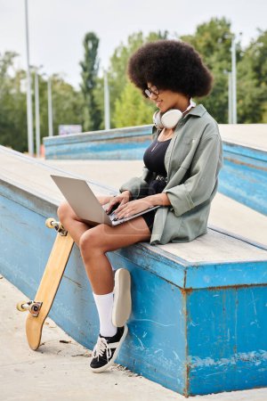 Eine junge Afroamerikanerin mit lockigem Haar sitzt auf einem Skateboard und tippt auf einem Laptop in einem Skatepark.