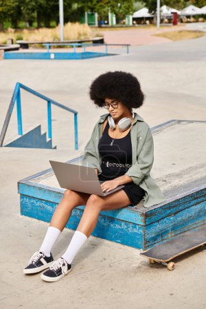 A young African American woman with curly hair seated on a skateboard, typing on a laptop in a skate park.