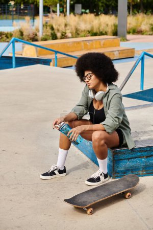 A young African American woman with curly hair sitting elegantly on a bench while holding a bottle of water in a skate park.