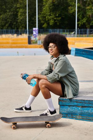 Junge Afroamerikanerin mit lockigem Haar sitzt auf einem Skateboard und hält eine Flasche Wasser in einem Outdoor-Skatepark.