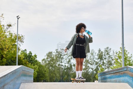 An African American woman with curly hair, standing on a skateboard, casually drinking water in a skate park.