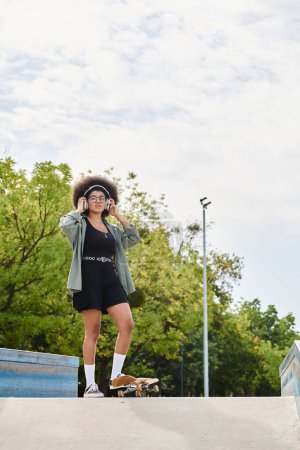 Foto de Una joven afroamericana con el pelo rizado se para con confianza en la parte superior de una rampa de skate en un parque de skate. - Imagen libre de derechos