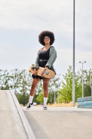 A young African American woman with curly hair confidently stands on a skateboard on a ramp in a skate park.