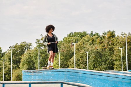 An African American woman with curly hair confidently stands on top of a skateboard ramp in an outdoor skate park.
