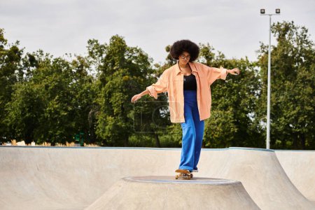 Foto de Una joven afroamericana con el pelo rizado monta con confianza su monopatín al lado de una rampa en un parque de skate. - Imagen libre de derechos