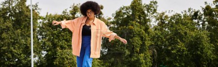 Téléchargez les photos : Une jeune afro-américaine aux cheveux bouclés se tient gracieusement sur une planche à roulettes, entourée d'arbres luxuriants dans un skate park. - en image libre de droit