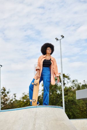 A young African American woman proudly holds a skateboard at the top of a ramp in an outdoor skate park.