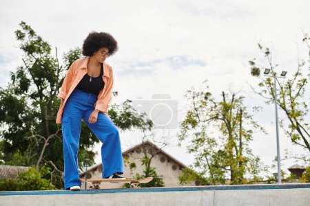Young African American woman with curly hair riding skateboard on top of cement wall in outdoor skate park.