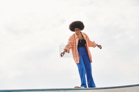 Eine junge Afroamerikanerin mit lockigem Haar steht selbstbewusst auf einem Skateboard an einer Rampe und zeigt ihre Skateboard-Fähigkeiten.