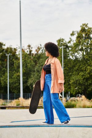 Young African American woman with curly hair walking casually in a skate park, holding a skateboard in her hand.