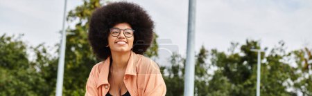 Téléchargez les photos : Une jeune femme afro-américaine élégante aux cheveux bouclés portant des lunettes et une chemise rose skateboard en plein air dans un skate park. - en image libre de droit