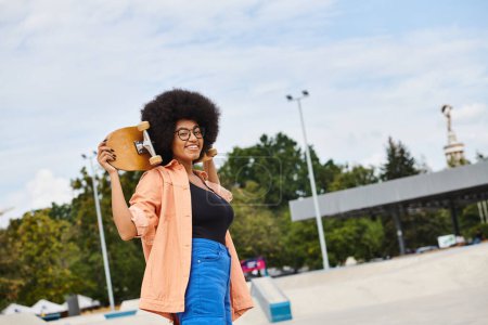 A stylish African American woman with curly hair holds a skateboard in her right hand at a skate park.