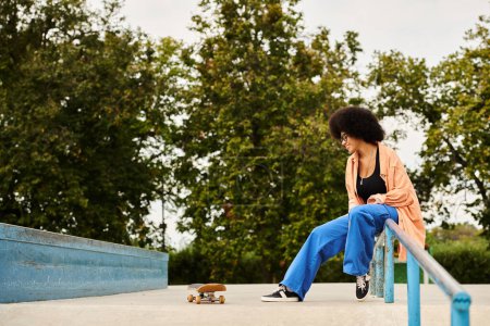 An African American woman with curly hair is sitting on a rail next to a skateboard in a skate park.