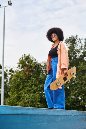 Junge Afroamerikanerin mit lockigem Haar hält selbstbewusst Skateboard, balanciert auf Rampe im Skatepark.