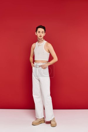 Photo for Stylish appealing gay man in casual attire with accessories posing on red backdrop and looking away - Royalty Free Image