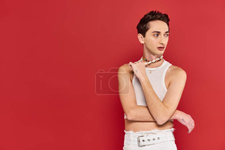 Photo for Attractive fashionable gay man in casual attire with accessories on red backdrop and looking away - Royalty Free Image