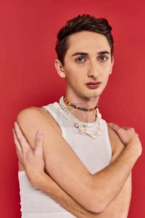 Photo for Appealing young gay man with stylish accessories in white attire looking at camera on red backdrop - Royalty Free Image