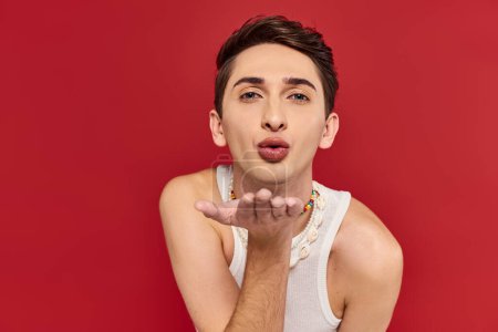 Téléchargez les photos : Attrayant gay l'homme en blanc casual tenue avec accessoires souffler baiser à caméra sur fond rouge - en image libre de droit
